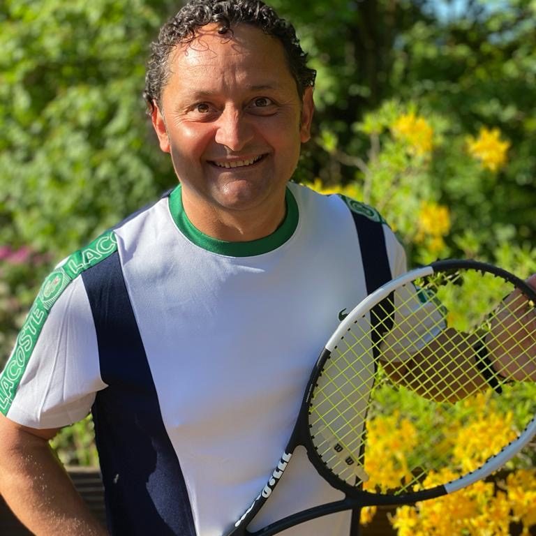 Mann hält einen Tennisschläger, lächelnd vor blühenden gelben Blumen.