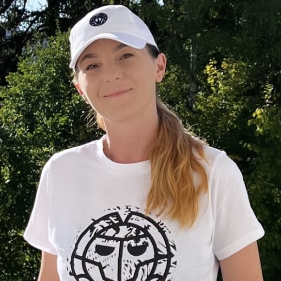 Frau mit langen, blonden Haaren, trägt ein weißes T-Shirt und eine schwarze Cap.