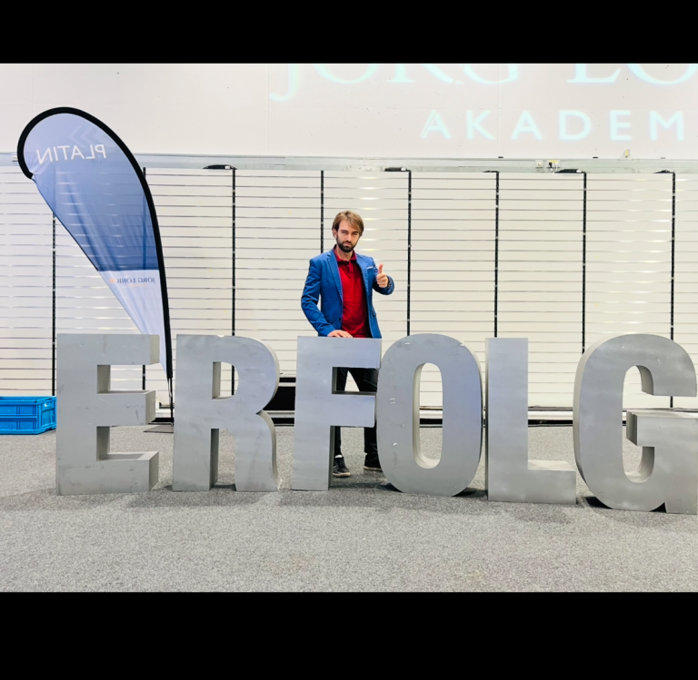 Mann in blauer Jacke steht neben großen Buchstaben "ERFOLG" bei einer Veranstaltung.