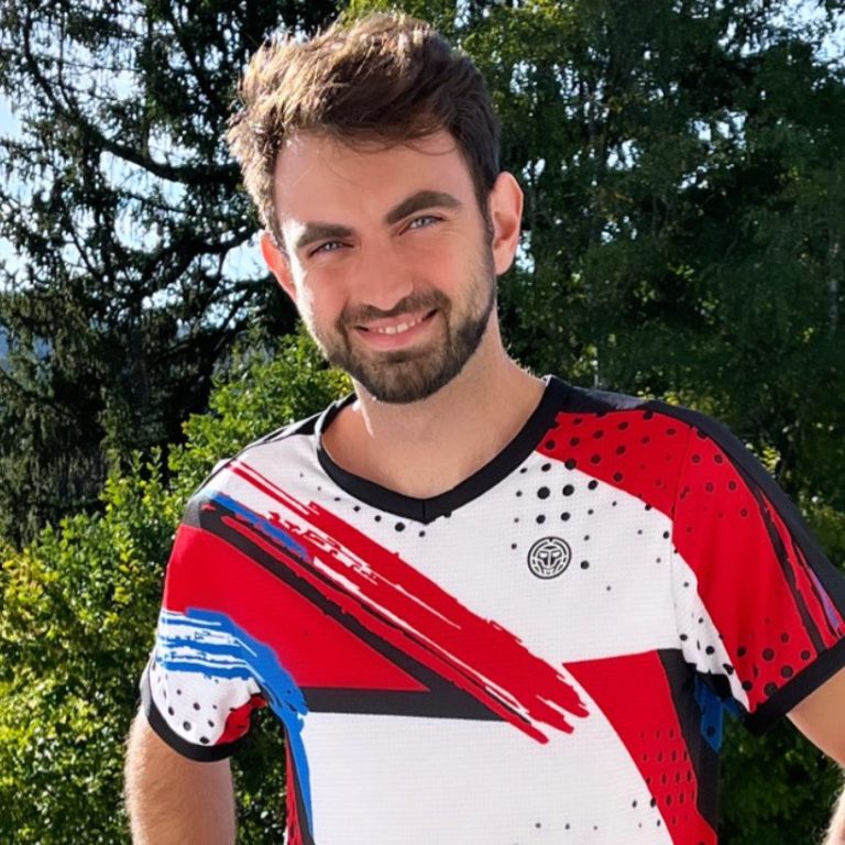Junger Mann in einem sportlichen Shirt mit bunten Mustern, lächelt vor grüner Kulisse.